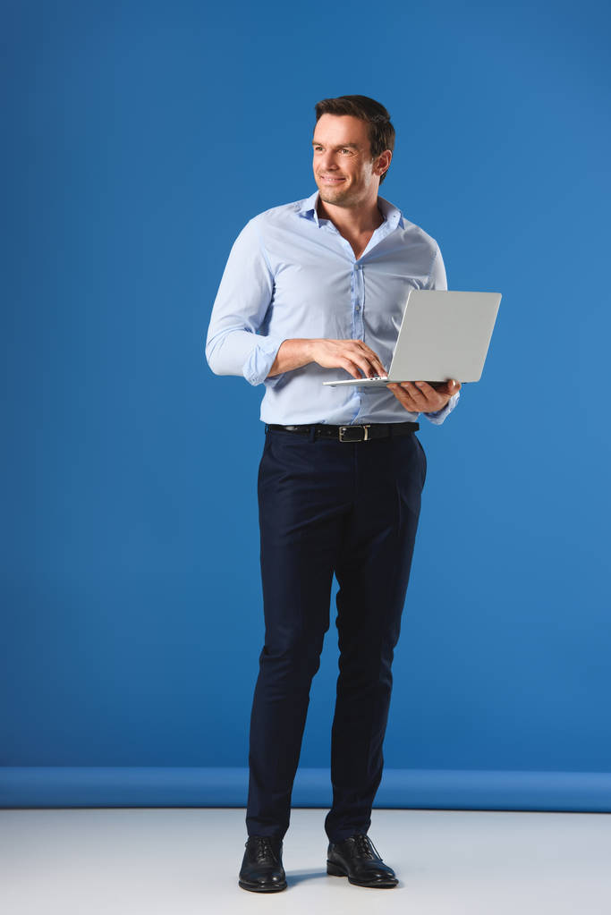 Вид в полный рост на красивого улыбающегося человека, держащего ноутбук и отводящего взгляд в сторону на голубом фоне
 - Фото, изображение