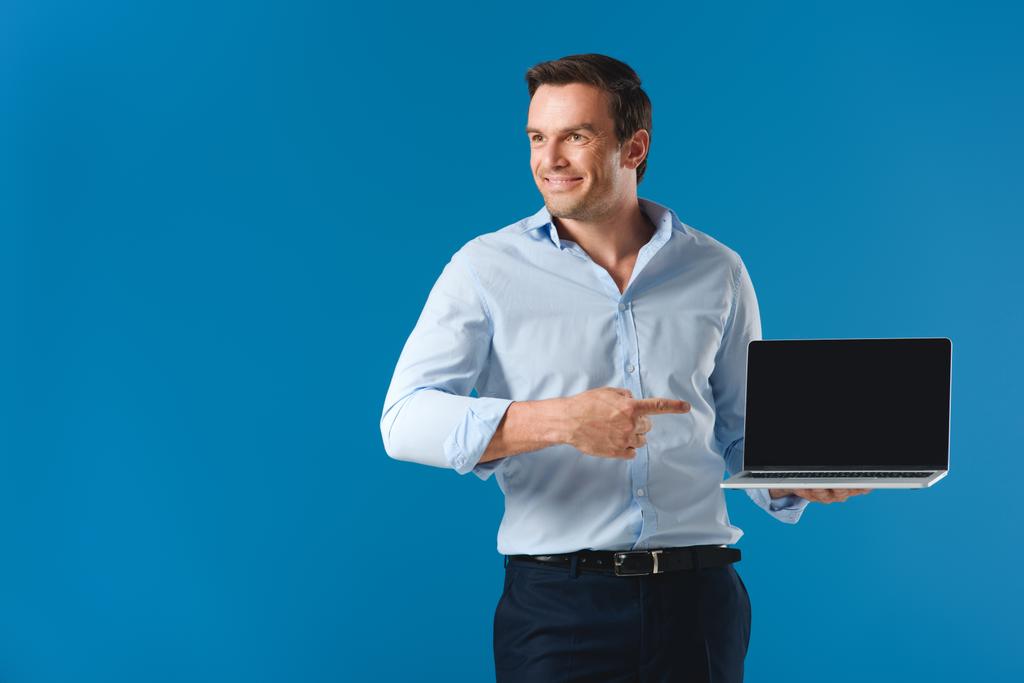 handsome smiling man pointing with finger at laptop with blank screen and looking away isolated on blue - Photo, Image