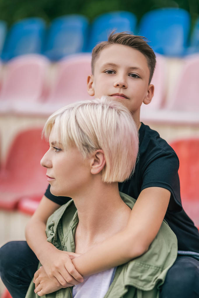 kaunis äiti ja poika syleilee istuessaan yhdessä stadionilla
 - Valokuva, kuva