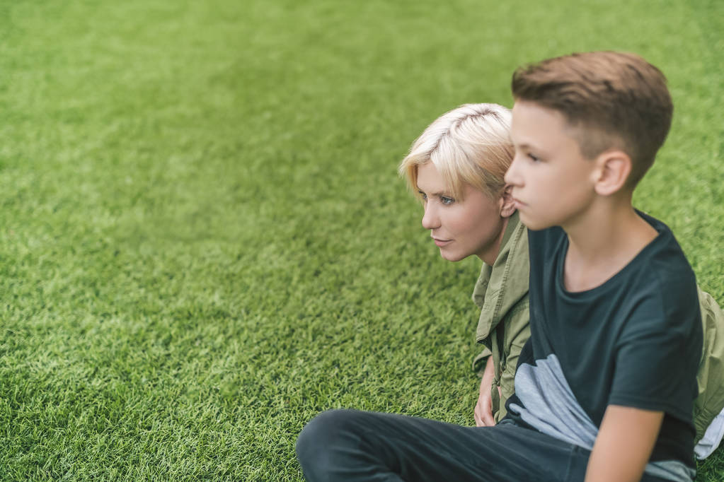 вид збоку красива мати і син дивиться в сторону, сидячи разом на траві
 - Фото, зображення