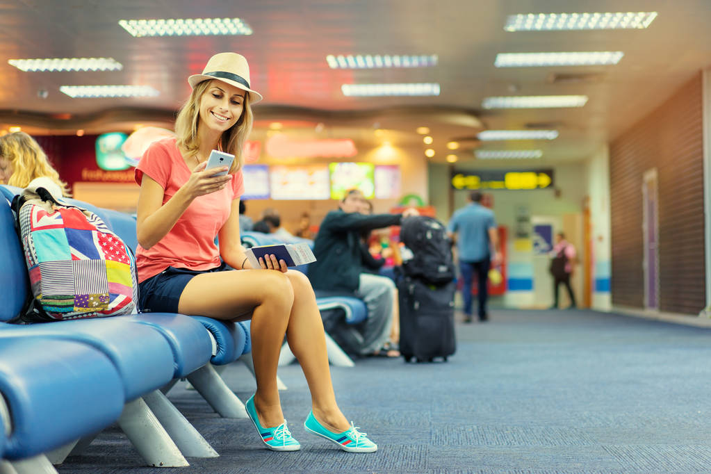 Красивая молодая женщина использует смартфон и держит билеты, сидя в терминале аэропорта в ожидании посадки
. - Фото, изображение