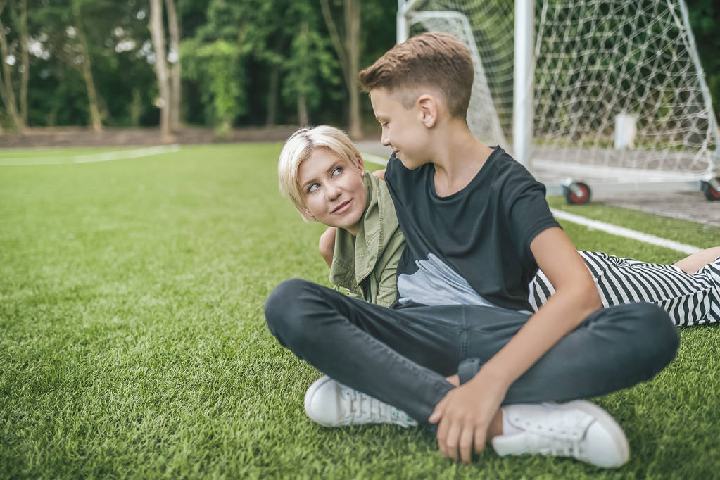 happy mother and son smiling each other while resting together on green lawn - Photo, Image