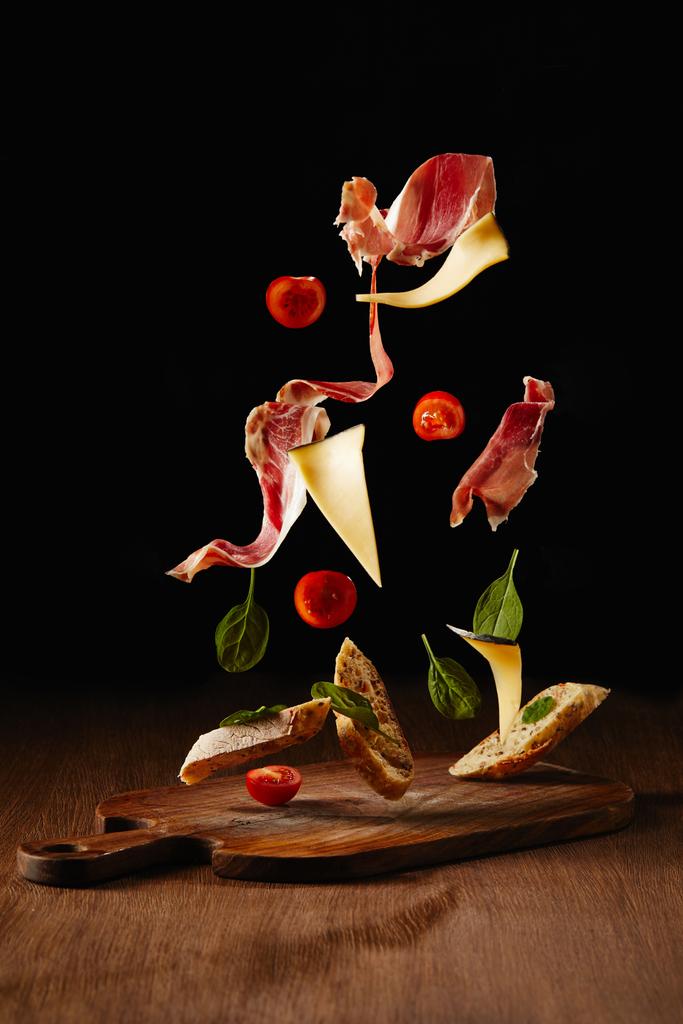 Ингредиенты для закусок с хлебом, хамоном и овощами, летящими над деревянной поверхностью стола
 - Фото, изображение