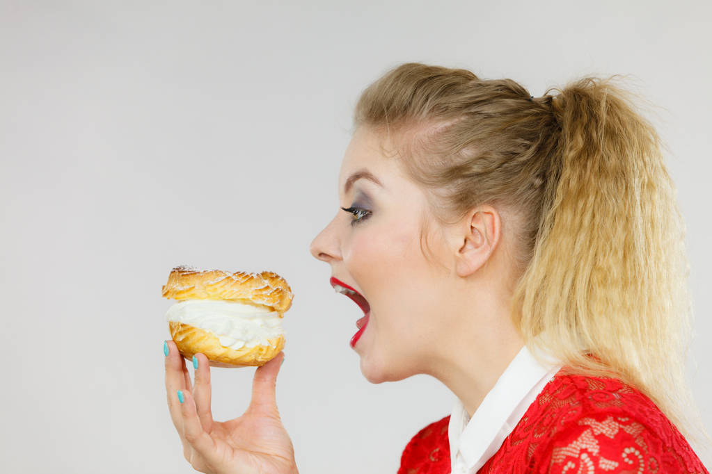 Słodkie koncepcji żywności i szczęścia. Zabawny radosny Kobieta blonde holding choux pyszne francuskie ciasto z bitą śmietaną, biorąc ogromny zgryz, podekscytowany wyrazem twarzy. Na szaro - Zdjęcie, obraz