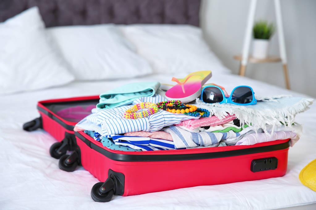 Avoin matkalaukku eri vaatteita ja asusteita kesämatkan sängyllä
 - Valokuva, kuva
