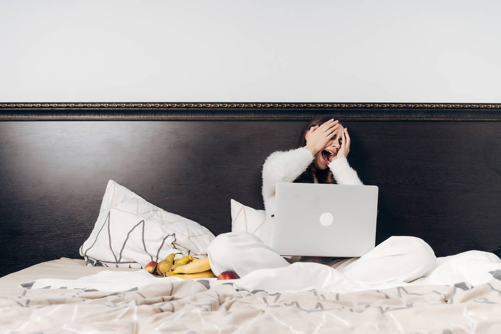 una chica en pijama blanco se sienta en la cama, viendo una película de miedo en su computadora portátil, cubriéndose la cara con las manos
 - Foto, imagen