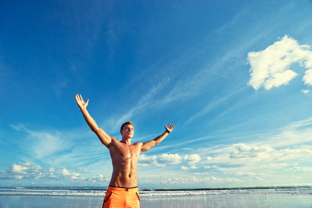 Young handsome strong man raising hands up on the beach against sky. - Photo, Image