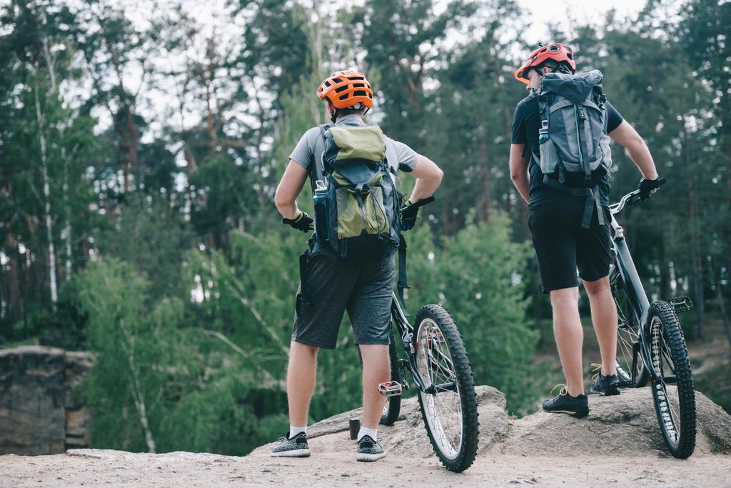 вид сзади на экстремальных велосипедистов-мужчин в защитных шлемах, стоящих с горными велосипедами в лесу
 - Фото, изображение