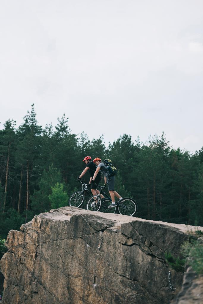 visão distante de ciclistas extremos masculinos em capacetes protetores andando em bicicletas de montanha em penhasco rochoso na floresta
 - Foto, Imagem