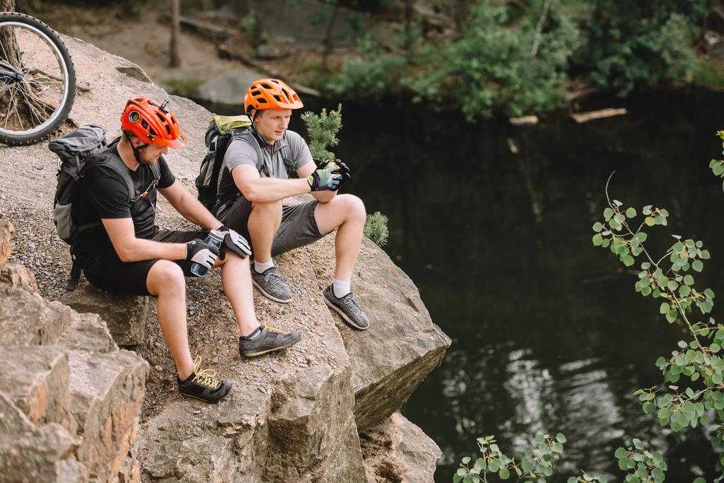 high angle view of friends cyclists with backpacks resting with sport bottle of water on rocky cliff near river in forest - Photo, Image