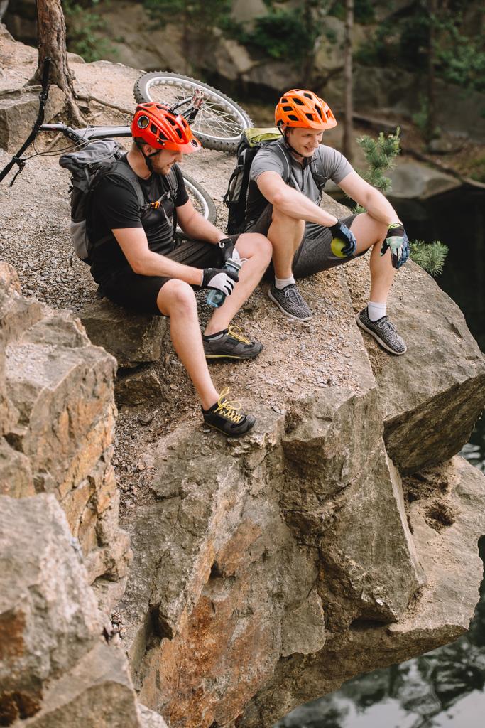 amis souriants cyclistes avec sacs à dos reposant avec bouteille d'eau de sport et pomme sur une falaise rocheuse près de la rivière en forêt
 - Photo, image