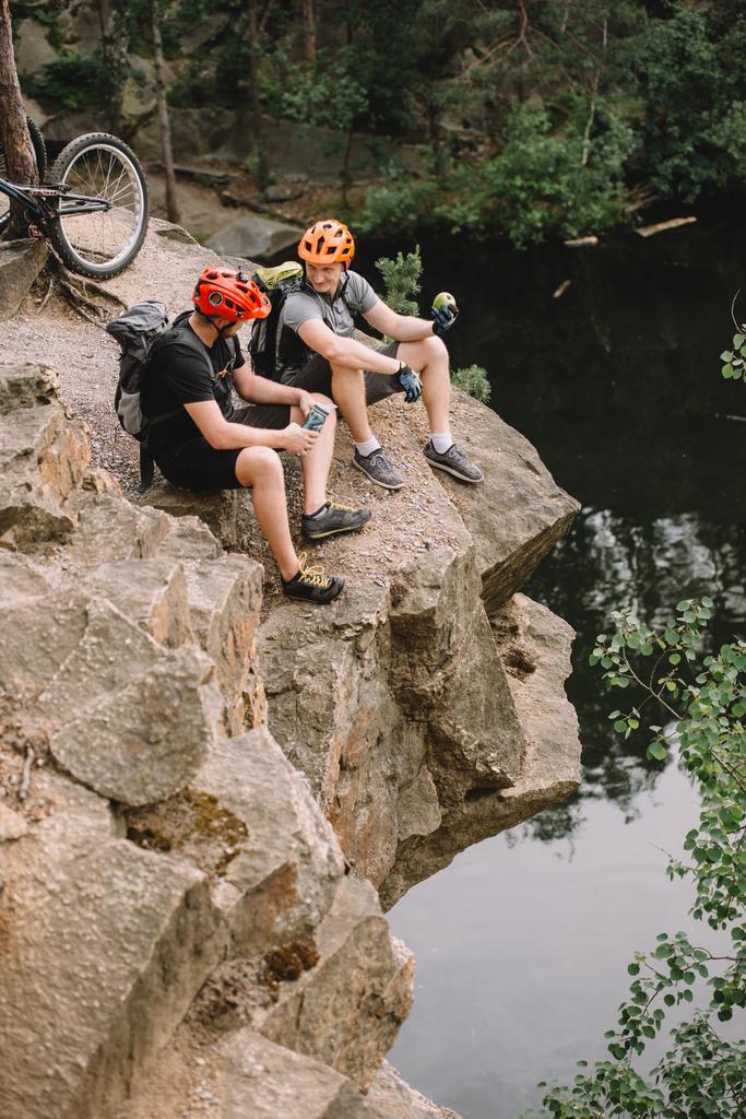 森の川の近くの岩の崖の水とリンゴのスポーツ ボトルと休憩のバックパックを友人の自転車のハイアングル - 写真・画像