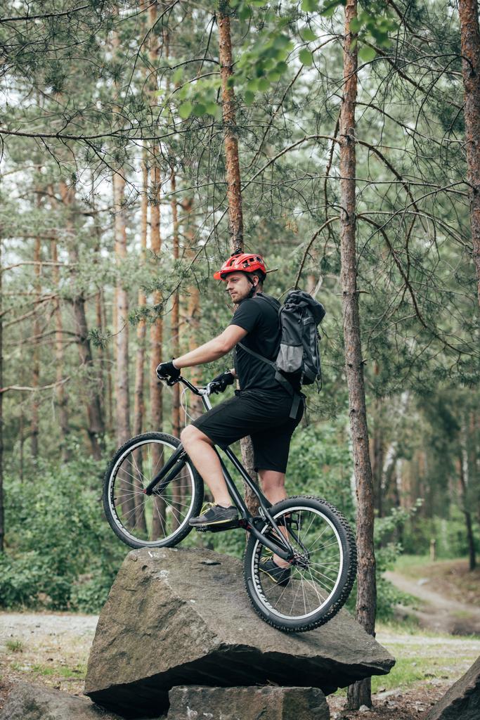 αρσενικό ακραία ποδηλάτη στο προστατευτικό κράνος στέκεται στο ποδήλατο βουνού σε πέτρα στο δάσος - Φωτογραφία, εικόνα