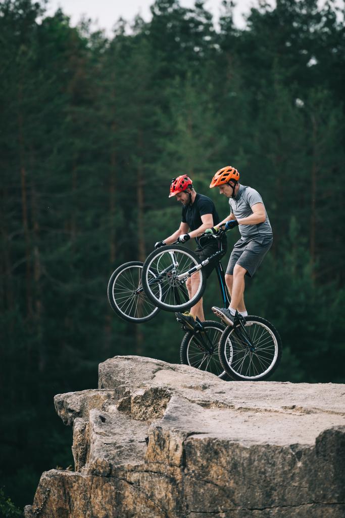 risky trial bikers standing on back wheels on rocky cliff with blurred pine forest on background - Photo, Image