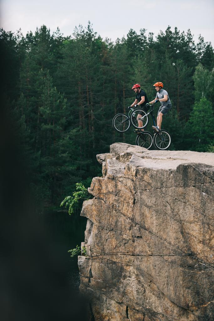 vista lateral de los ciclistas de trial de pie sobre ruedas traseras en acantilado rocoso con bosque de pino borroso en el fondo
 - Foto, Imagen