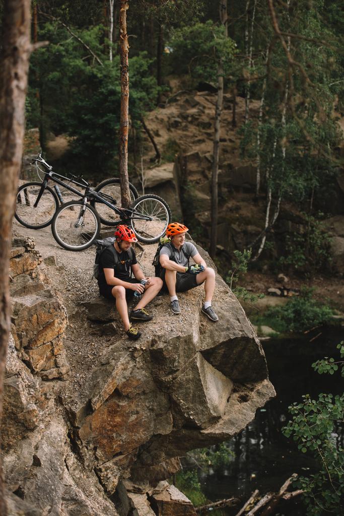 vista de ángulo alto de los jóvenes ciclistas de trial felices relajándose en el acantilado rocoso después del paseo
 - Foto, imagen