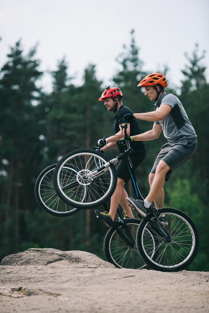 przystojny młodych rowerzystów próbny stojąc na tylne koła na skalistym klifie z niewyraźne sosnowego lasu na tle - Zdjęcie, obraz