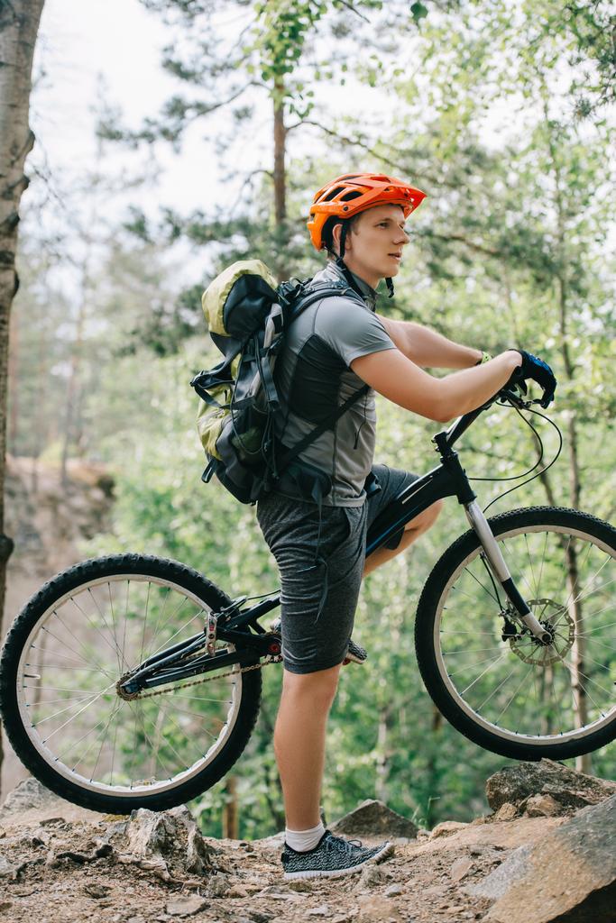 Seitenansicht eines Radfahrers mit Helm und Rucksack, der mit Mountainbike im Wald steht  - Foto, Bild