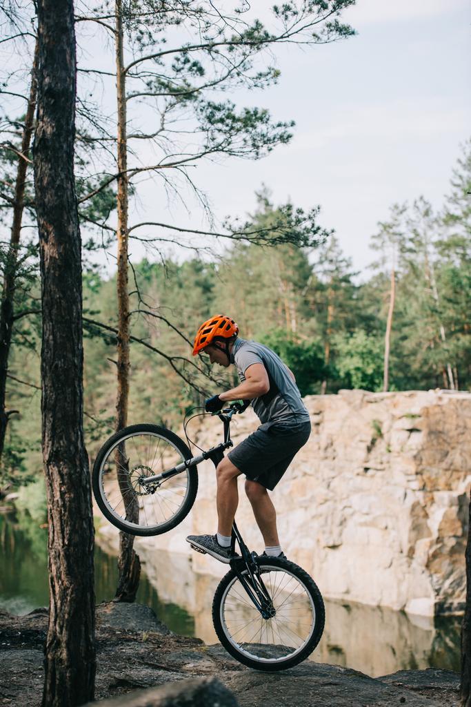 Seitenansicht des Extremradfahrers im Schutzhelm balanciert auf dem Hinterrad des Mountainbikes im Wald - Foto, Bild
