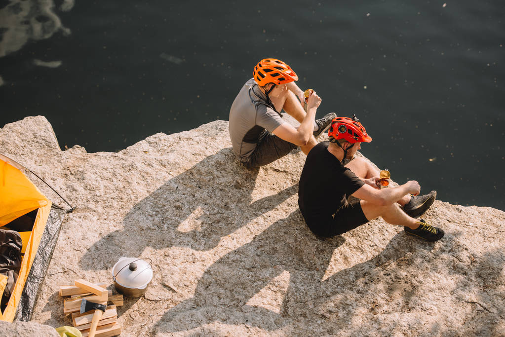 viajantes em capacetes de proteção comer alimentos enlatados perto de tenda, logs, machado e caldeirão em penhasco rochoso sobre o rio
  - Foto, Imagem