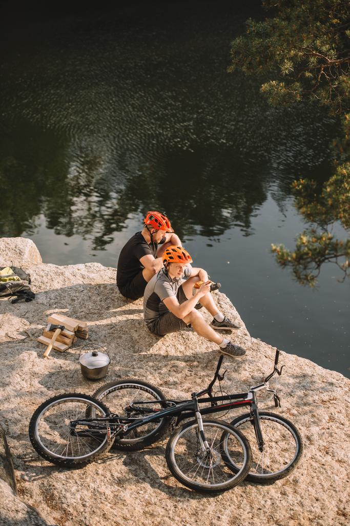 dwóch rowerzystów próbny odpoczynku w pobliżu dzienniki, kocioł i góry cykli na skalistym urwisku nad rzeką - Zdjęcie, obraz