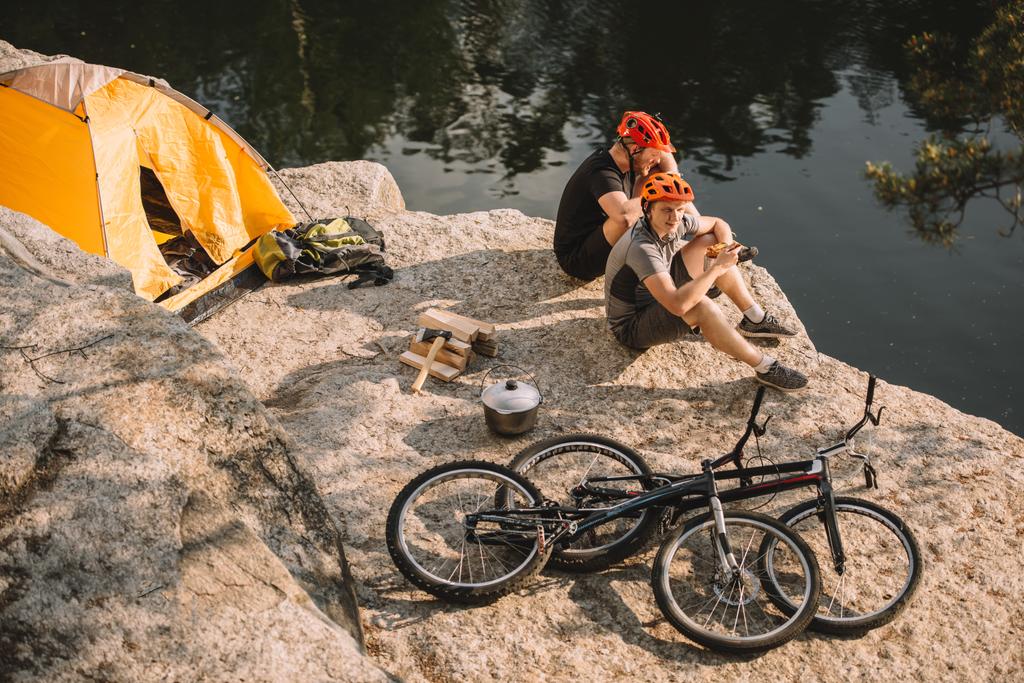 ciclistas de trial descansando cerca de tienda y ciclos en acantilado rocoso sobre el río
 - Foto, Imagen