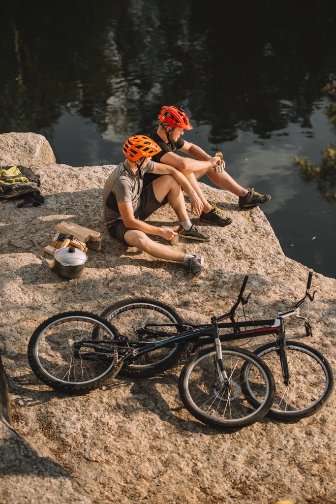 high angle view of trial bikers resting near logs, cauldron and mountain cycles on rocky cliff over river - Photo, Image