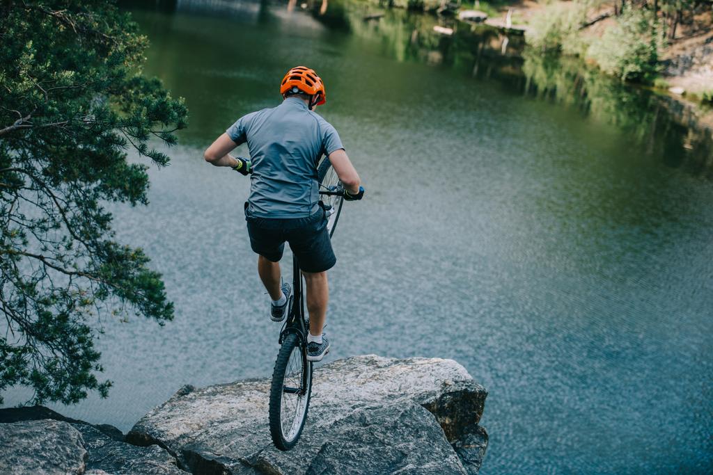 young trial biker balancing on back wheel on rocky cliff over lake - Photo, Image