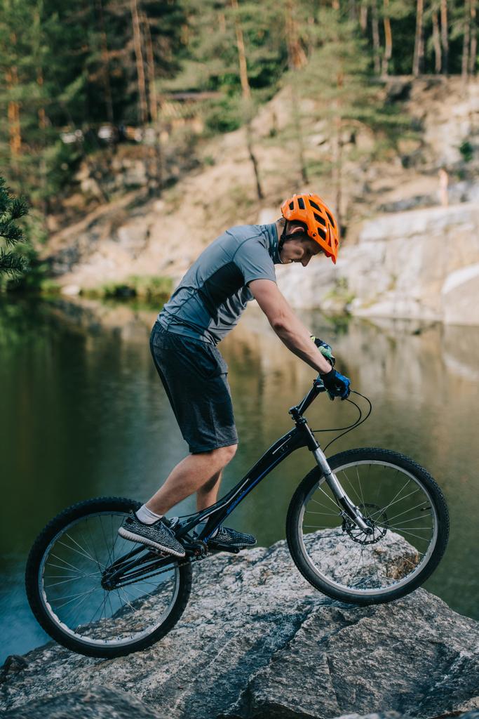 νεαρός δοκιμή ποδηλάτης έλεγχοι εξισορρόπησης σε βραχώδη πλαγιά, πάνω στη λίμνη - Φωτογραφία, εικόνα