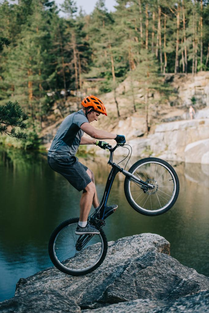 Sportlicher Trial-Biker balanciert auf Hinterrad an Felswand über See - Foto, Bild