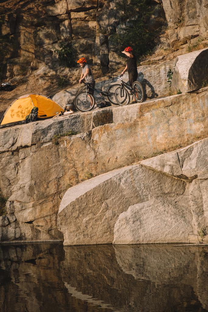 川に岩の崖にあるテントの近くのマウンテン バイクで立っている 2 つの男性方の遠景 - 写真・画像