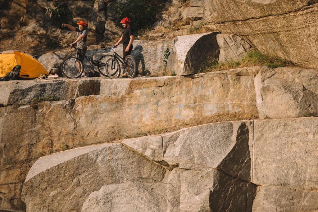 viajante masculino em capacete de proteção em pé com mountain bike e apontando por dedo para amigo perto da barraca em penhasco rochoso
  - Foto, Imagem
