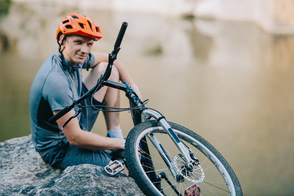 усміхнений молодий пробний велосипедист розслабляється на скелястій скелі над водою і дивиться на камеру
 - Фото, зображення