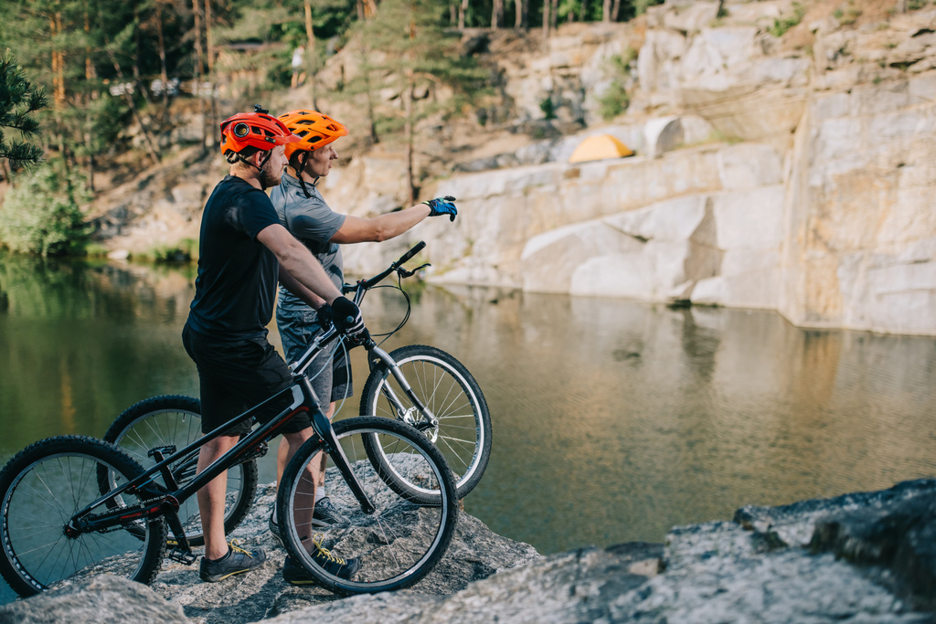 Junge Trial-Biker stehen auf Felswand vor Bergsee und zeigen auf Seite - Foto, Bild