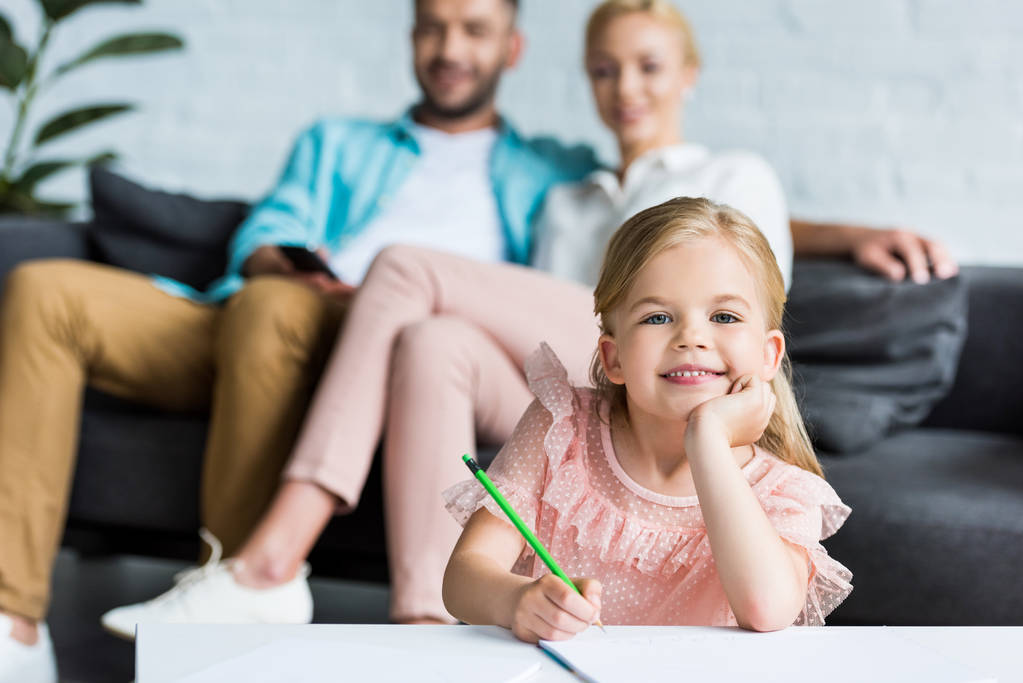 adorable child drawing with pencil and smiling at camera while parents sitting behind - Photo, Image