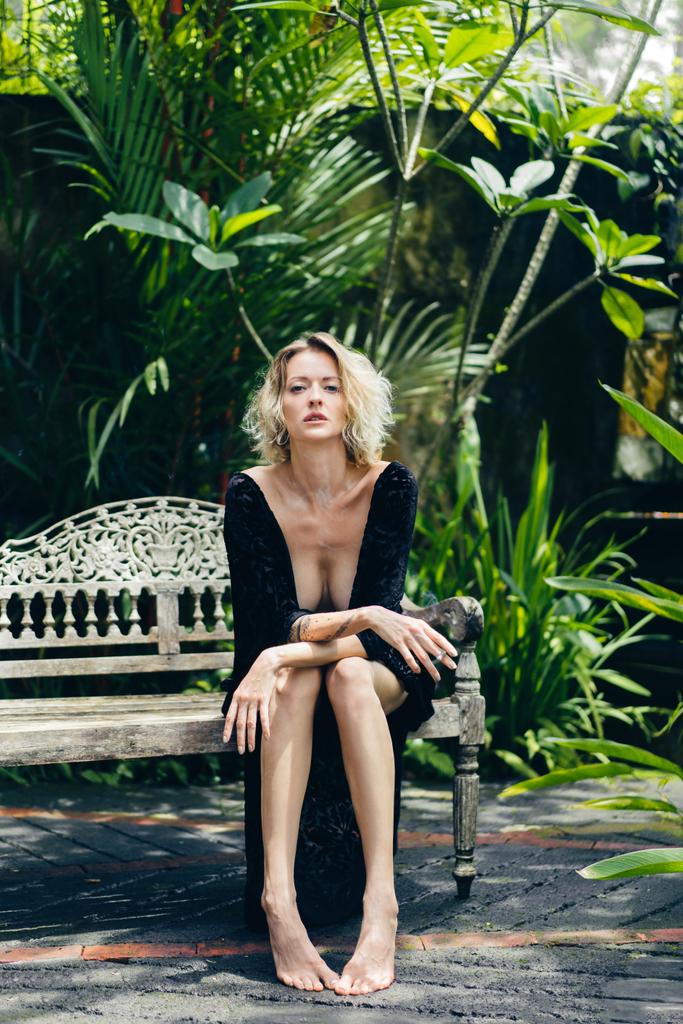 blond woman in black clothing with cigarette in hand resting on bench on terrace, ubud, bali, indonesia - Photo, Image
