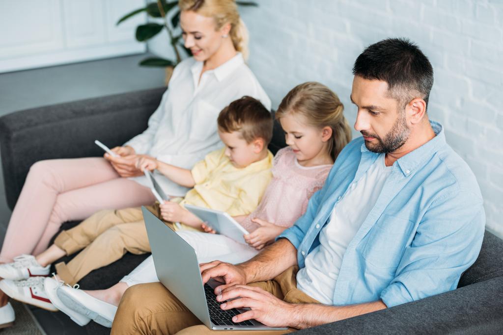 perhe, jossa kaksi lasta istuu sohvalla ja käyttää digitaalisia laitteita kotona
  - Valokuva, kuva