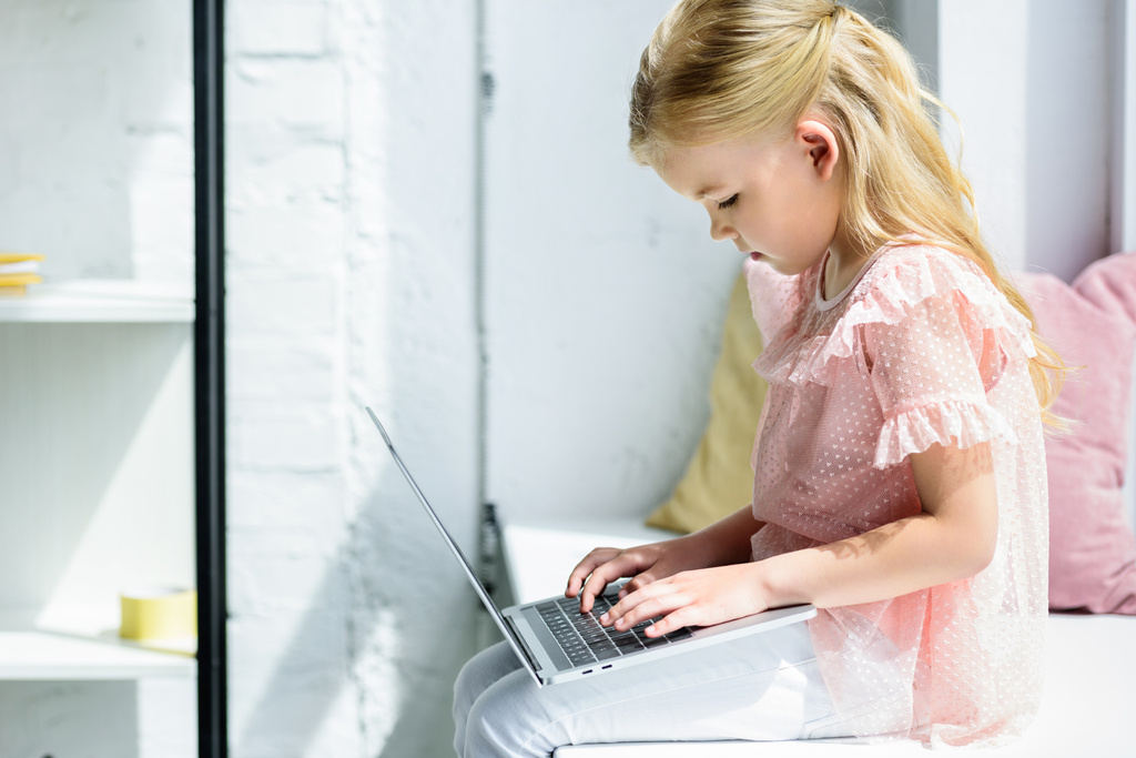 Seitenansicht des niedlichen kleinen Kindes mit Laptop auf der Fensterbank - Foto, Bild