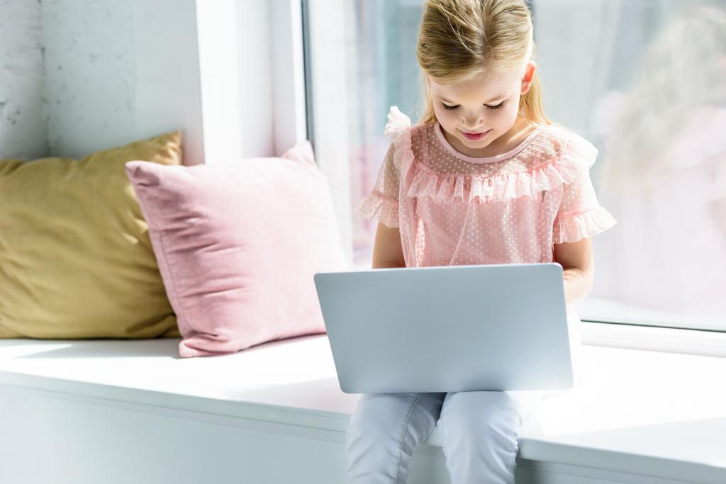 αξιολάτρευτο μικρό παιδί κάθεται στο περβάζι και χρησιμοποιούν φορητό υπολογιστή - Φωτογραφία, εικόνα