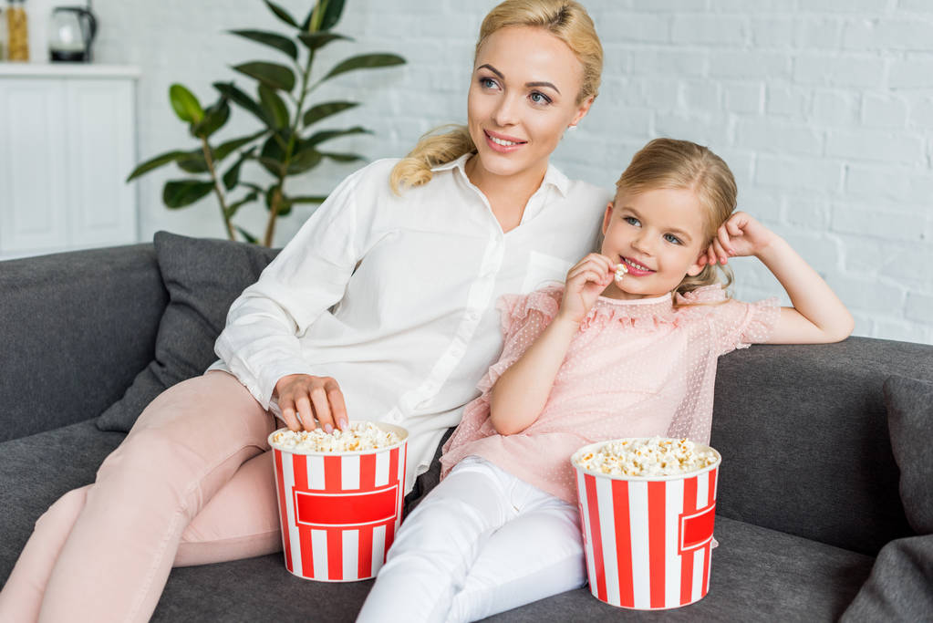 piękny uśmiechający się matka i córka jedzenia popcorn i odwracając w domu  - Zdjęcie, obraz