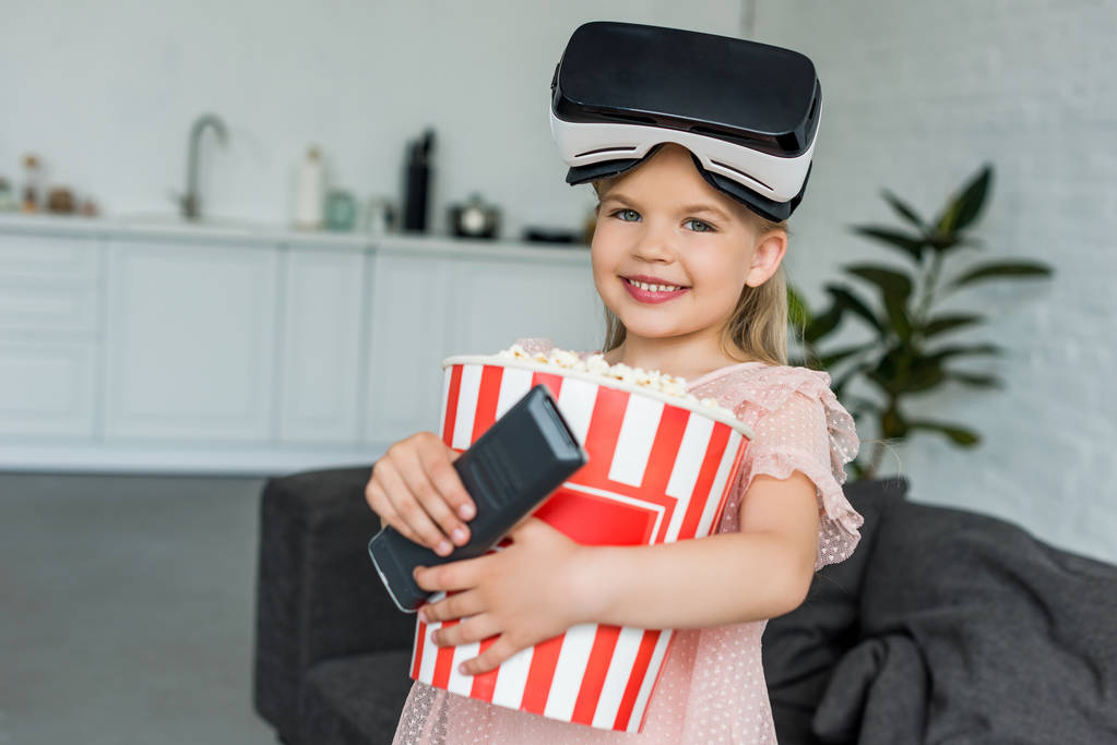 rozkošný usmívající se dítě v držení box s popcorn a vzdálený řadič soupravu pro virtuální realitu - Fotografie, Obrázek