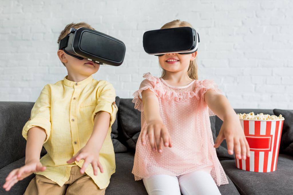 Imádnivaló kis gyerekek virtuális valóság fejhallgatók használata közben ül a kanapén, otthon - Fotó, kép