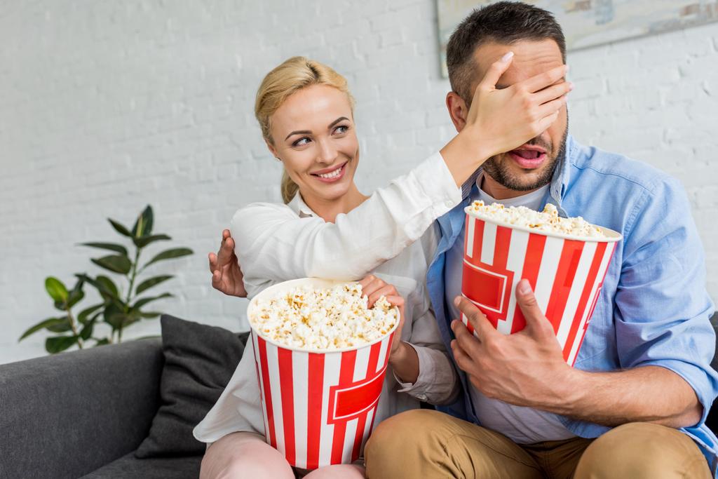 smiling woman closing eyes to husband while eating popcorn and watching tv at home - Photo, Image