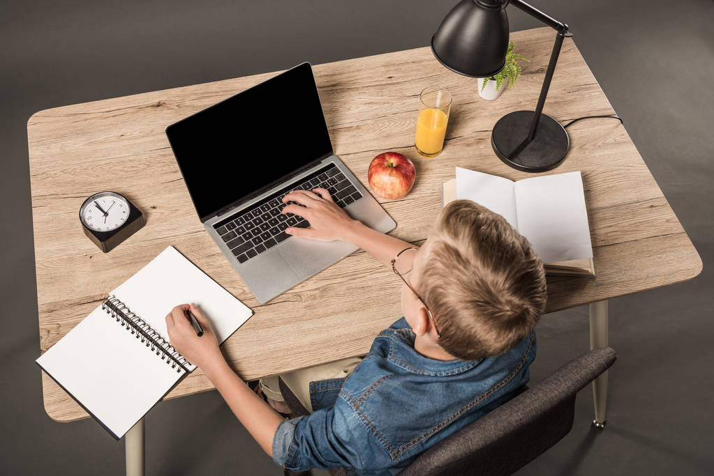 vue surélevée de l'écolier faisant ses devoirs sur ordinateur portable à la table avec manuel, livre, plante, lampe, horloge, pomme et verre de jus
  - Photo, image