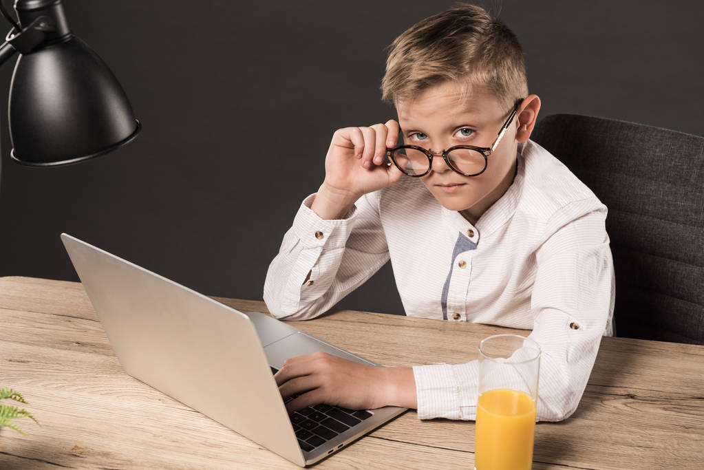 серйозний маленький хлопчик в окулярах дивиться на камеру, сидячи за столом з ноутбуком, лампою і склянкою соку на сірому фоні
  - Фото, зображення