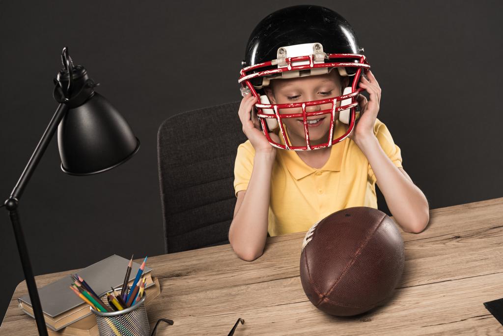 colegial sonriente que se pone el casco de fútbol americano en la mesa con bola, lámpara, lápices de color y libros sobre fondo gris
  - Foto, Imagen