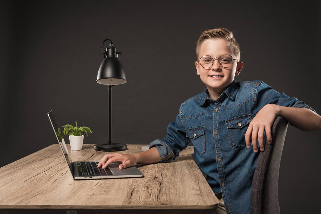 glimlachend jongetje in brillen met behulp van laptop aan tafel met plant en lamp op grijze achtergrond  - Foto, afbeelding