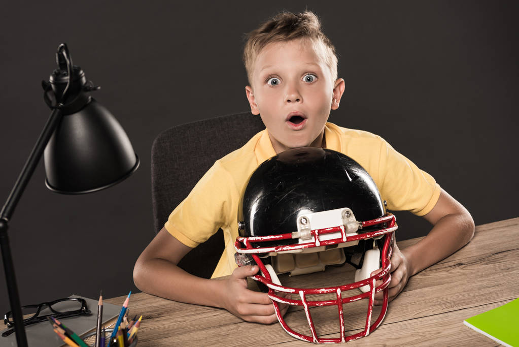 shocked schoolboy sitting with american football helmet at table with eyeglasses, lamp, colour pencils and books on grey background  - Photo, Image