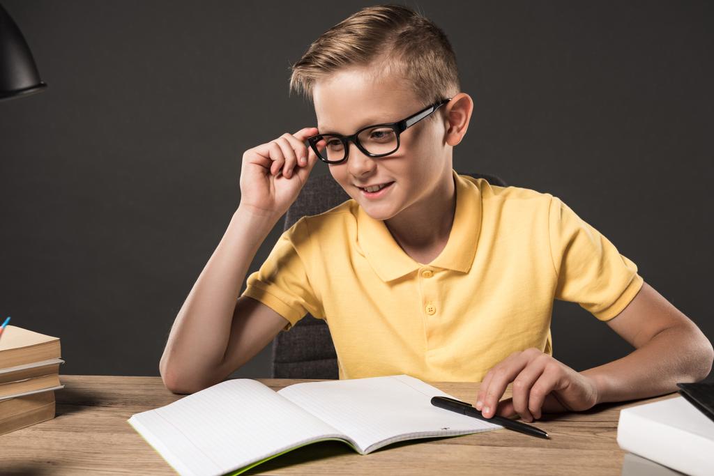 書籍、教科書、灰色の背景の上にランプのスタックでテーブルで宿題をしながらよそ見眼鏡の男子生徒の笑顔  - 写真・画像