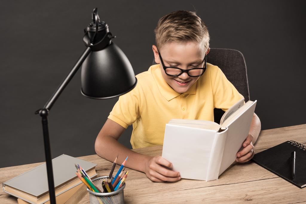 μαθητής σε γυαλιά ανάγνωσης βιβλίου στο τραπέζι με χρώμα μολύβια, βιβλία, συγγράμματα και λαμπτήρα σε γκρίζο φόντο  - Φωτογραφία, εικόνα
